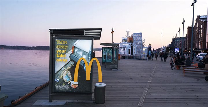 McDonalds-campagneposter met de tekst 'Take away your take away' op een bushokje. De M uit het logo verbindt de poster met een afvalbak naast de abri.