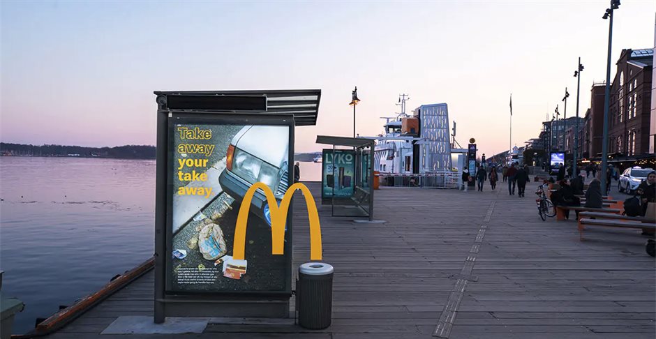 McDonalds-campagneposter met de tekst 'Take away your take away' op een bushokje. De M uit het logo verbindt de poster met een afvalbak naast de abri.